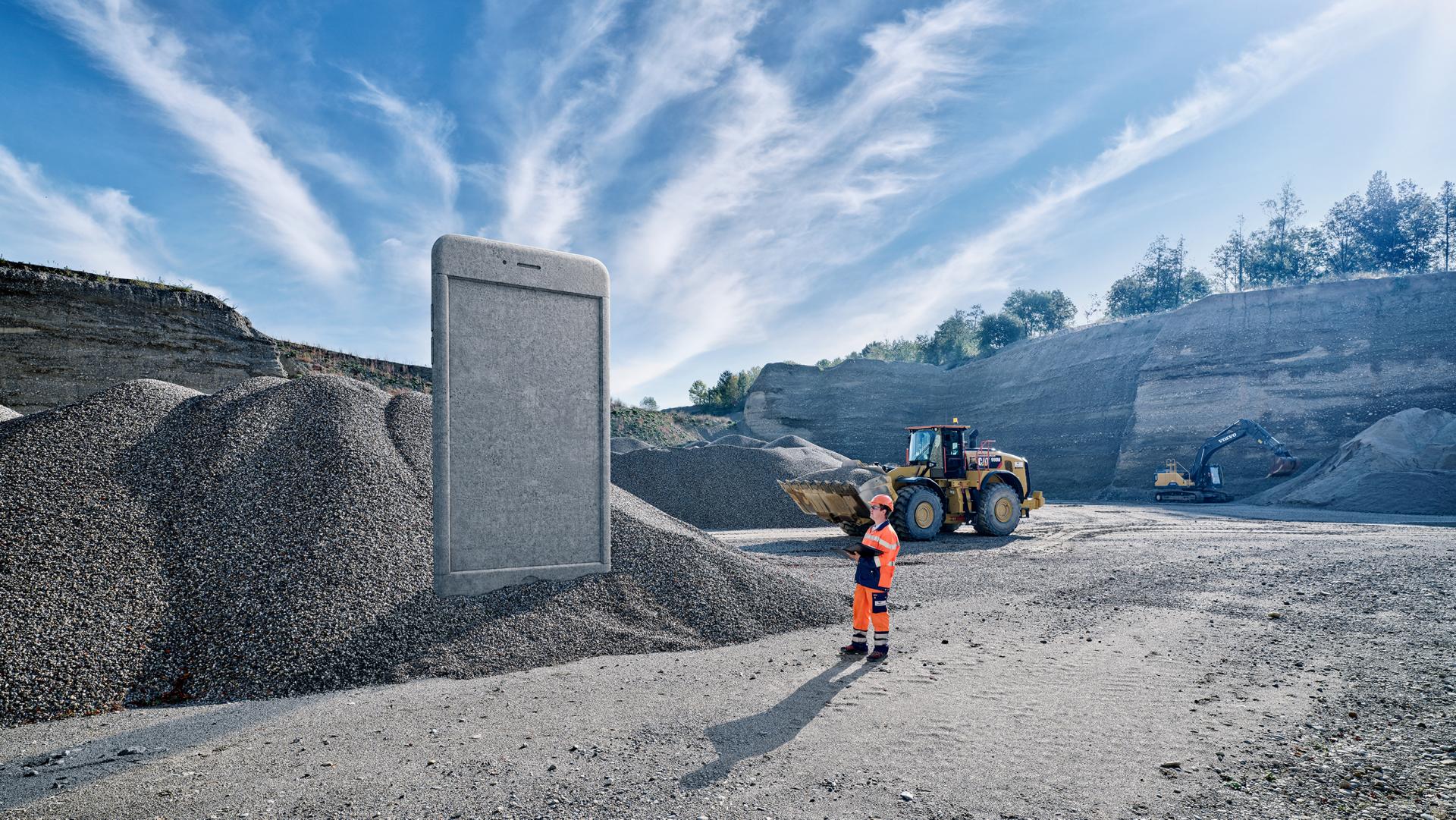 Vigier Beton Keyvisual: In einer Kiesgrube schaut sich ein Mitarbeiter ein riesiges Beton-Smartphone an