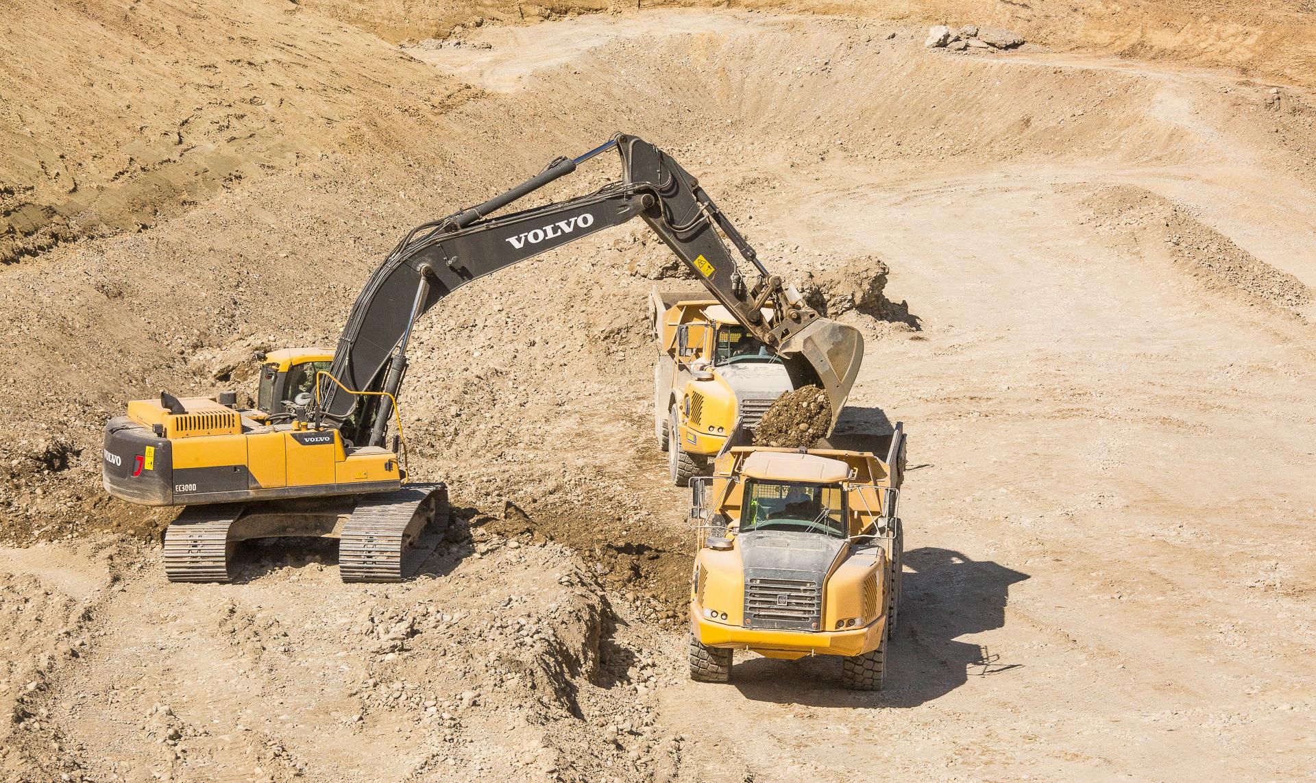 Ein gelber Bagger befüllt eine Mulde auf einem Transporter. Sie stehen in einer Grube.