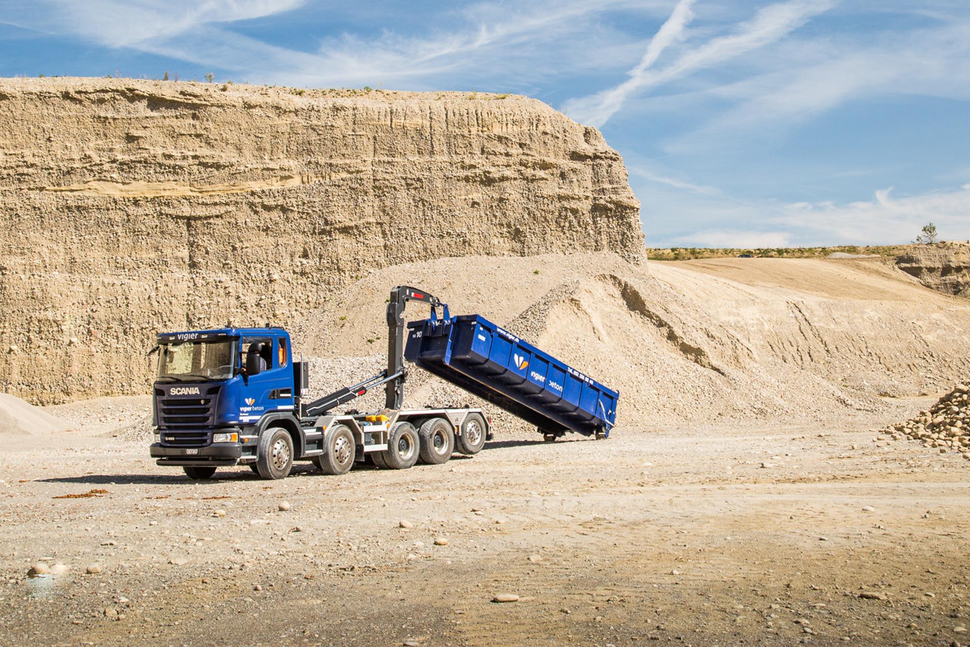 Ein Lastwagen entlädt eine grosse, blaue Mulde in Mitten eines Steinbruchs.
